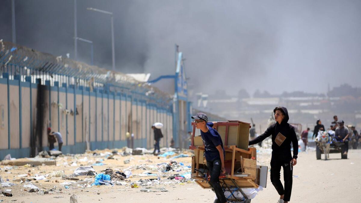 فلسطينيون يفرون بأمتعتهم بينما يتصاعد الدخان في الخلفية، في منطقة تل السلطان في رفح.  – الصورة: وكالة فرانس برس